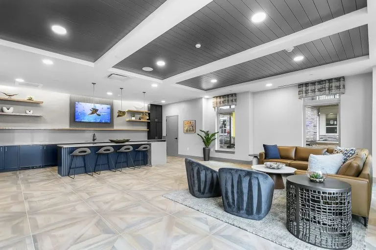 Community lounge with a wall-mounted TV, bar seating, and blue armchairs around a rug and coffee tables